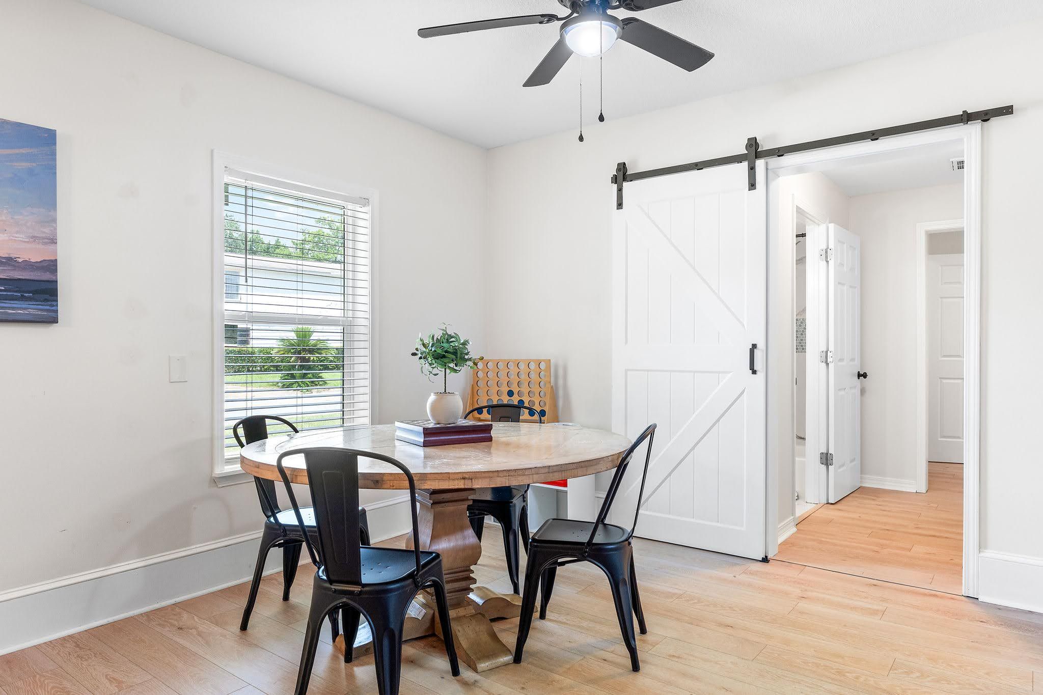Un coin repas comprenant une table ronde, quatre chaises, un ventilateur de plafond et une vue sur l’extérieur par une fenêtre.