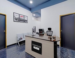 A reception desk with a sign reading 'TOWNHOUSE' and 'OYO-Serviced'. There are framed pictures on the wall, a computer, and a chair with a bag. - Classic MJR Square Mall (Hyderabad)