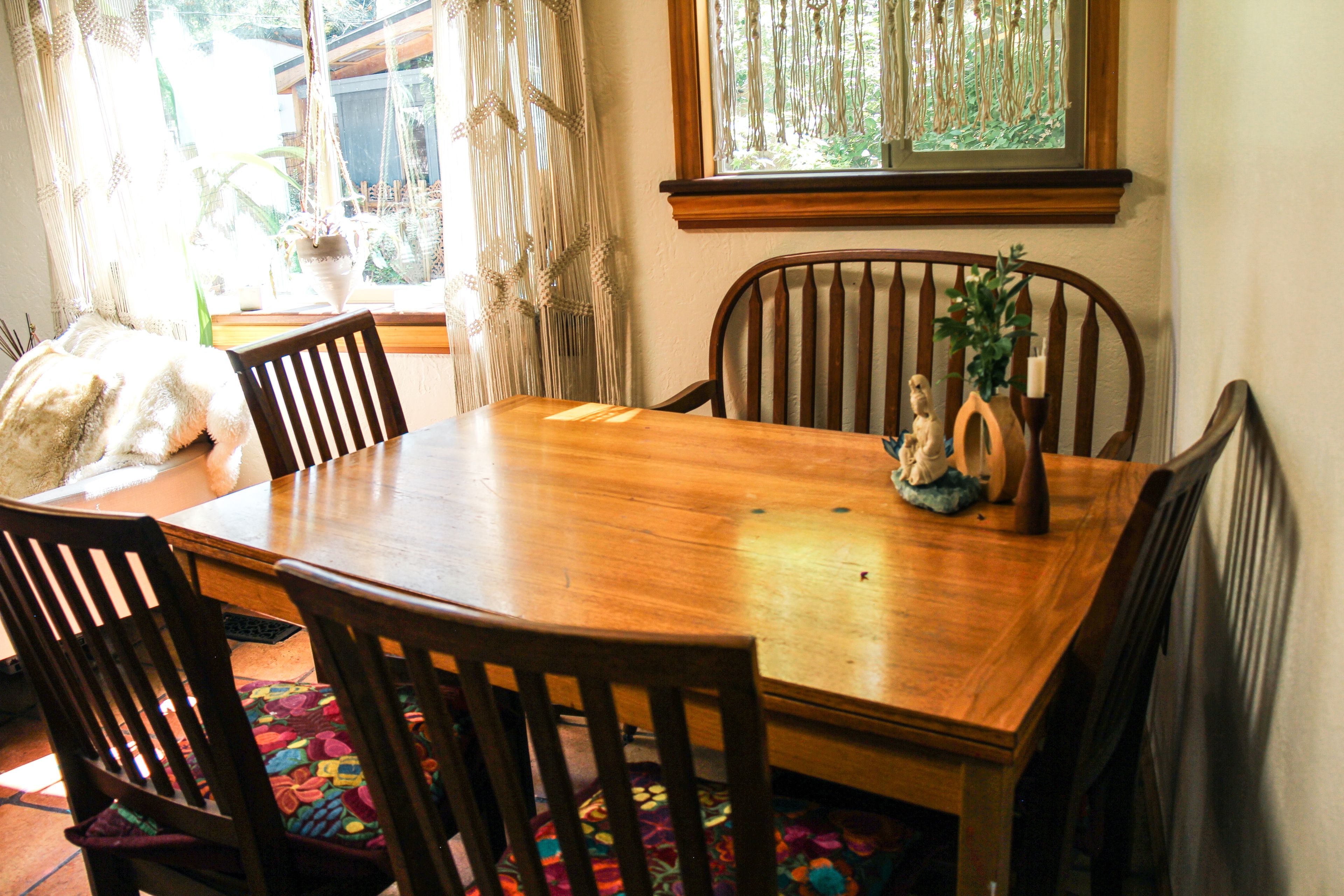 Una mesa de comedor de madera con seis sillas, una planta decorativa y una ventana con cortinas de macramé.