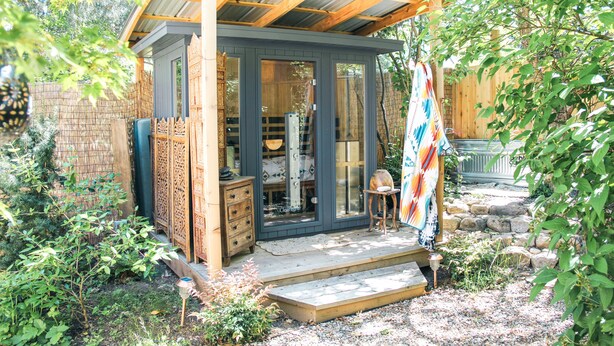 Una piccola struttura in legno con tetto metallico, una porta in vetro e un gradino che conduce a una zona con posti a sedere.