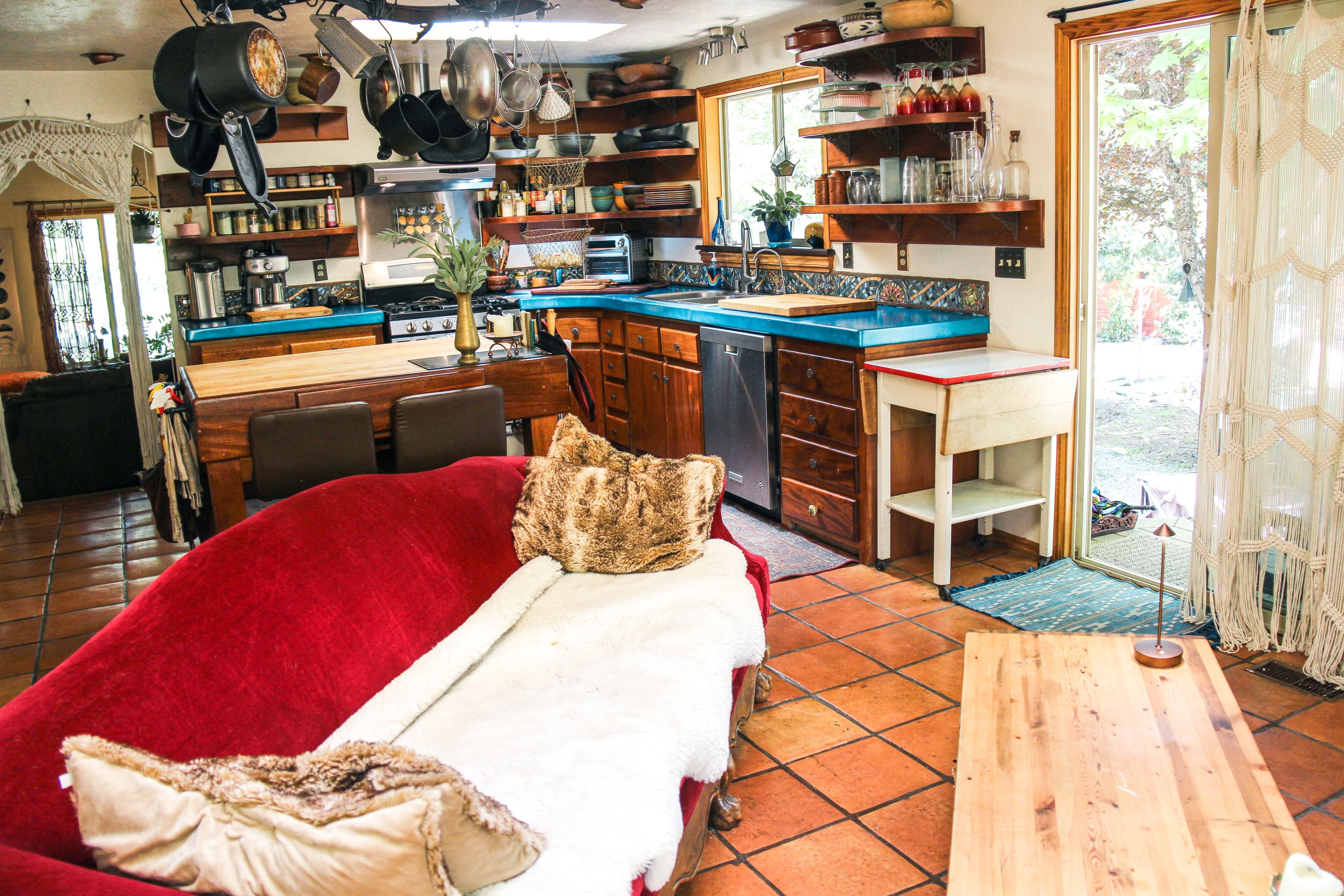 Een gezellige woonkamer met een rode bank, een houten eettafel en een keuken met blauwe aanrechtbladen en diverse keukengerei.