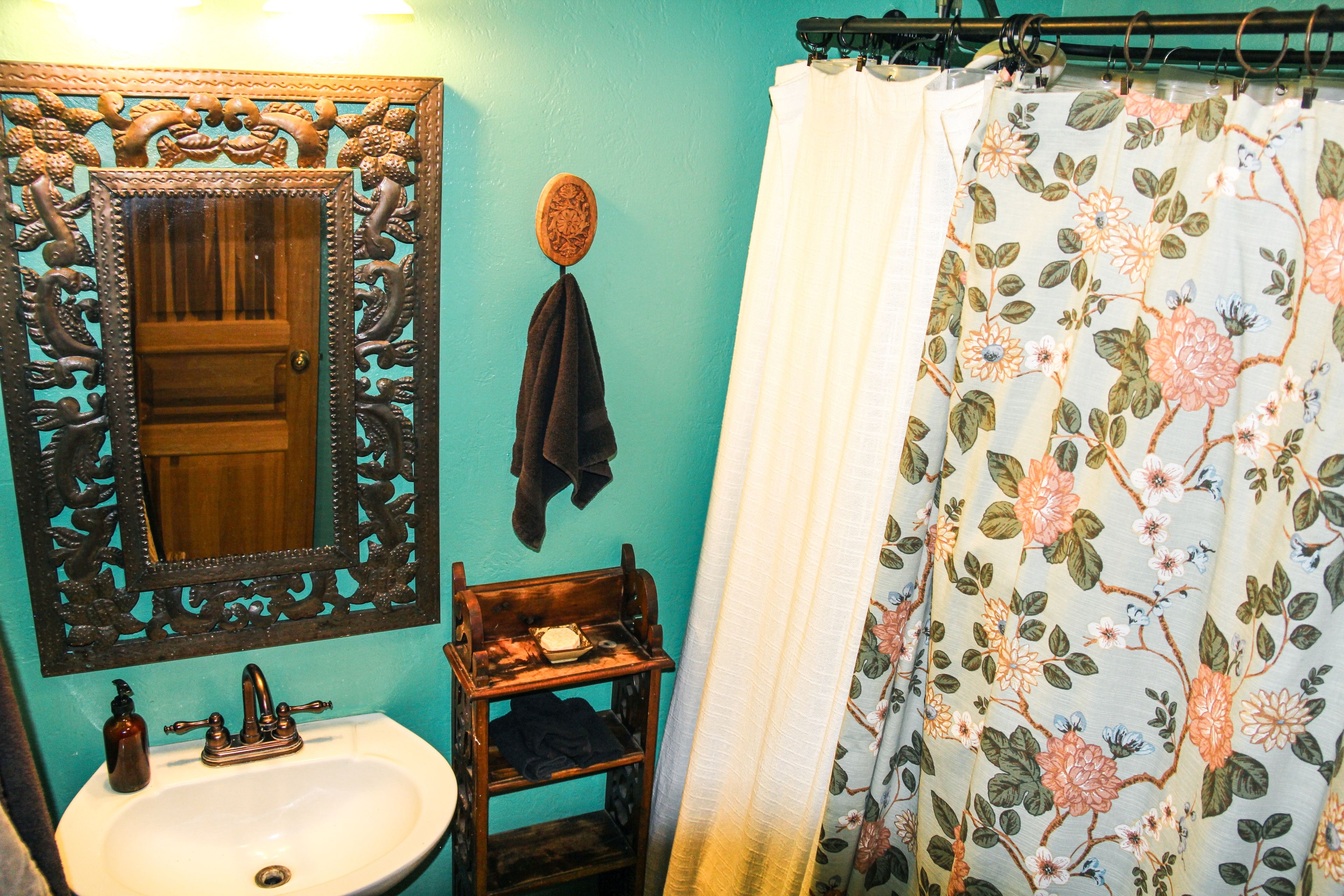 A bathroom with a wooden vanity, a sink, a mirror with ornate carvings, a towel hanging on a hook, and a floral shower curtain.