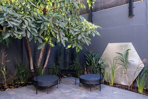 An outdoor seating area with two chairs and a table, surrounded by various plants and a large geometric wall structure.