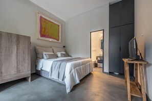 A modern bedroom with a large bed, a wardrobe, a desk with a computer, and a bathroom in the background.