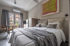 Ein Hotelzimmer mit Bett, Schreibtisch und Stuhl. Es gibt ein Fenster mit Vorhängen, eine Wand-Klimaanlage und ein Bild an der Wand.