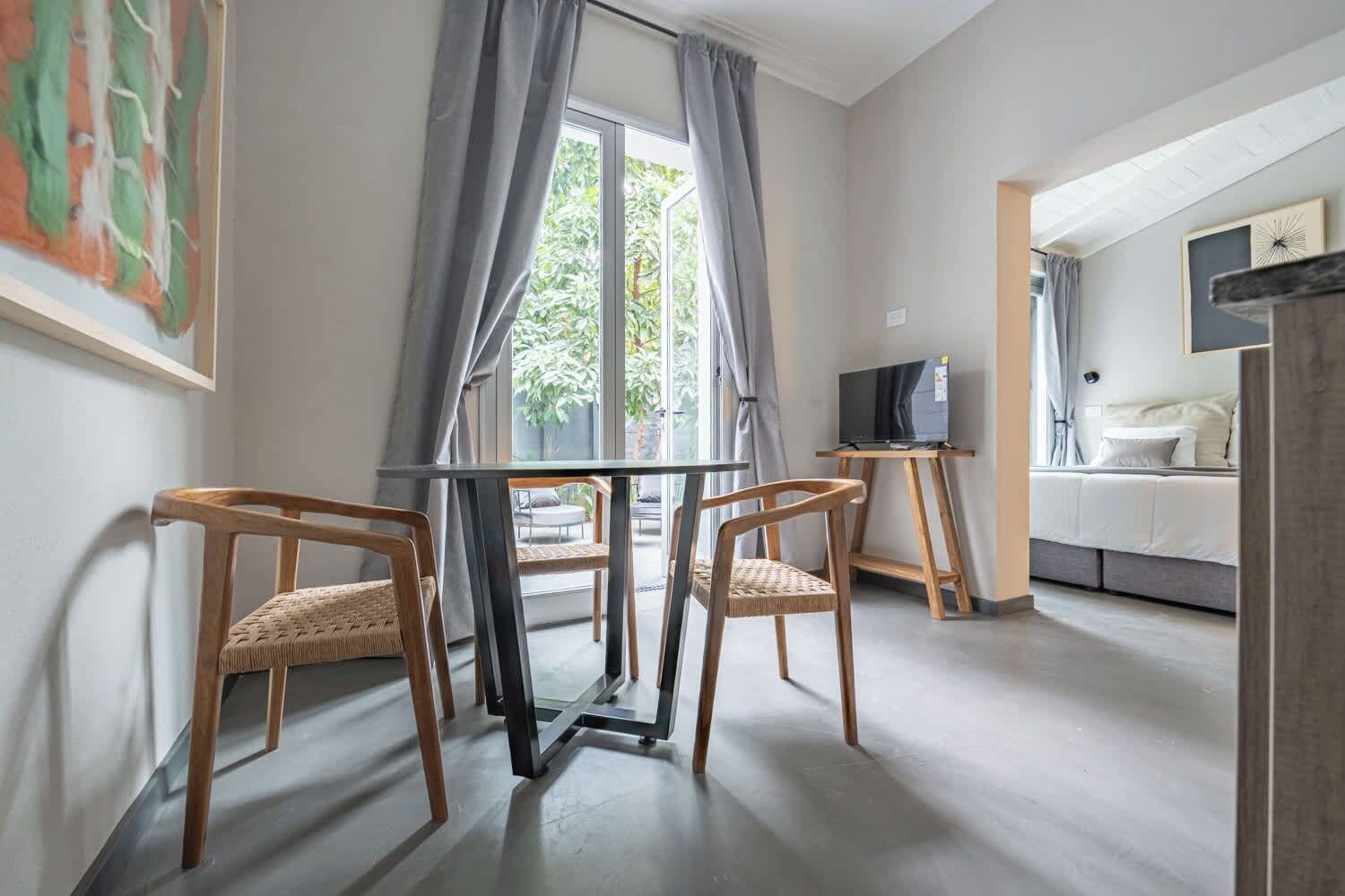 A room with a dining table, two chairs, a TV on a stand, and a bed with a headboard.