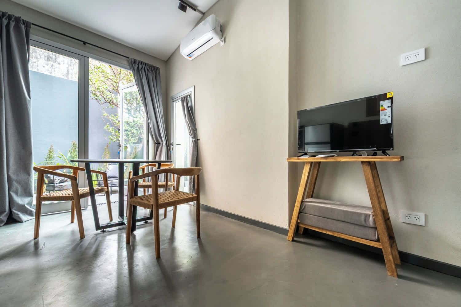 Una habitación con televisión sobre un mueble de madera, una mesa con sillas y ventanas grandes con cortinas.