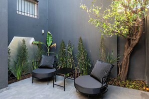 Un pequeño espacio al aire libre con dos sillas y una mesa, rodeado de plantas y un árbol.