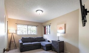 A room with a sectional sofa, a small cabinet, and a lamp. There are two windows with curtains, and a ceiling light fixture. - Summerlin Woods Paradise — Your Perfect Florida Getaway! (Fort Myers)