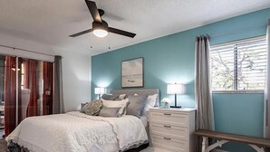 Quarto com cama, mesas de cabeceira, ventoinha no teto e janela com cortinas.