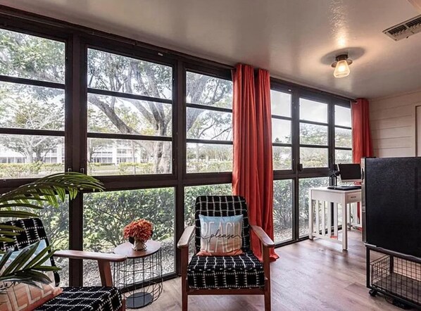 Une pièce avec de grandes fenêtres, un fauteuil avec un coussin, une petite table avec un vase et un meuble pour la télévision.