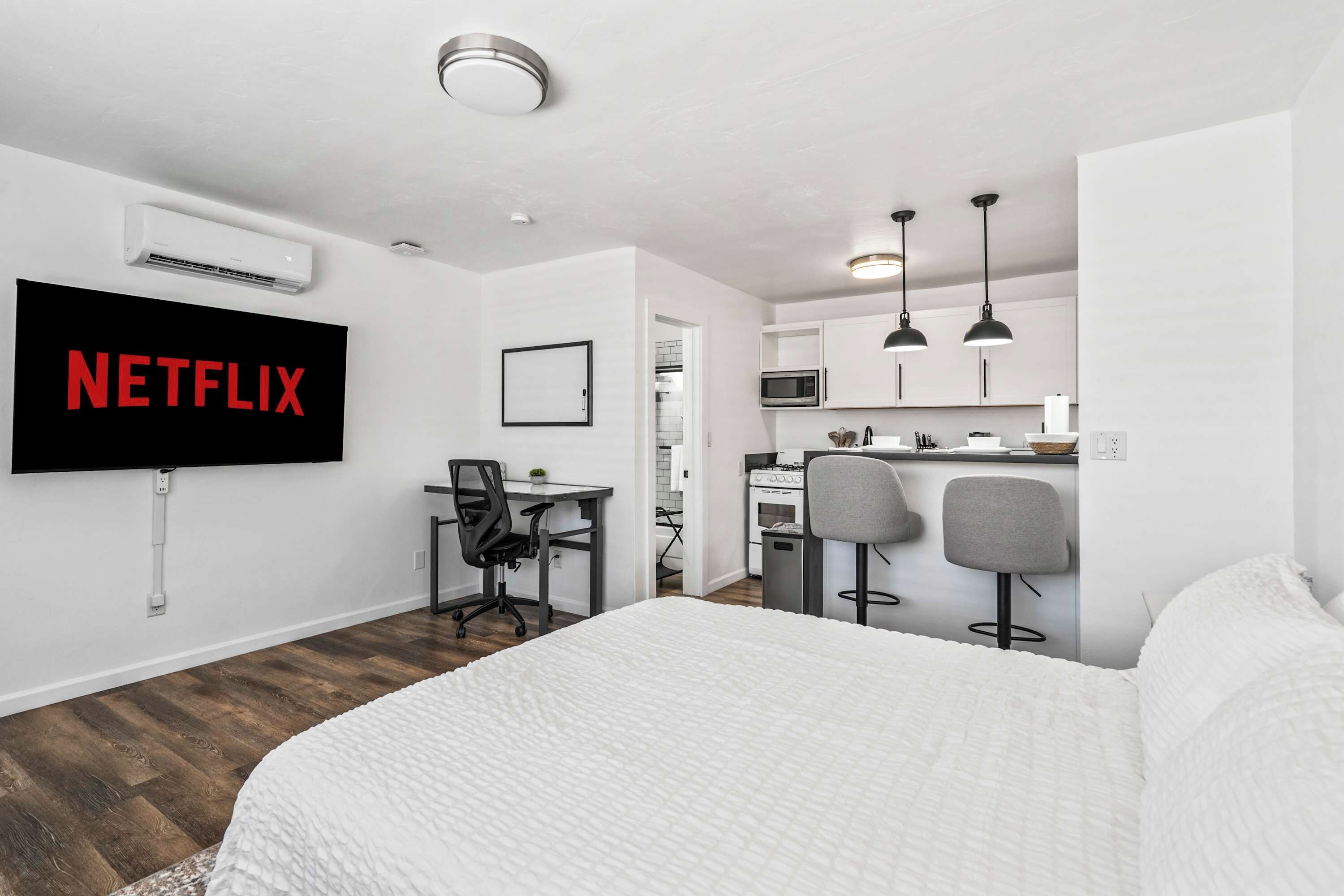 A modern hotel room with a bed, a desk, a chair, a TV mounted on the wall, and a kitchenette.