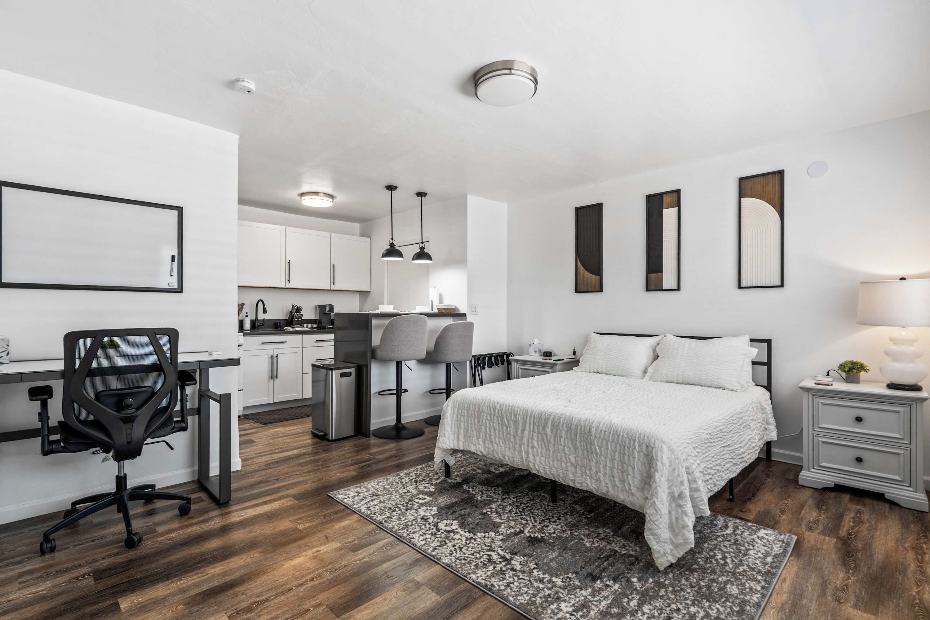 A modern bedroom with a bed, bedside tables, a desk with a chair, and a kitchen area in the background.
