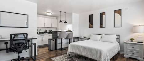 A modern bedroom with a bed, bedside tables, a desk with a chair, and a kitchen area in the background.