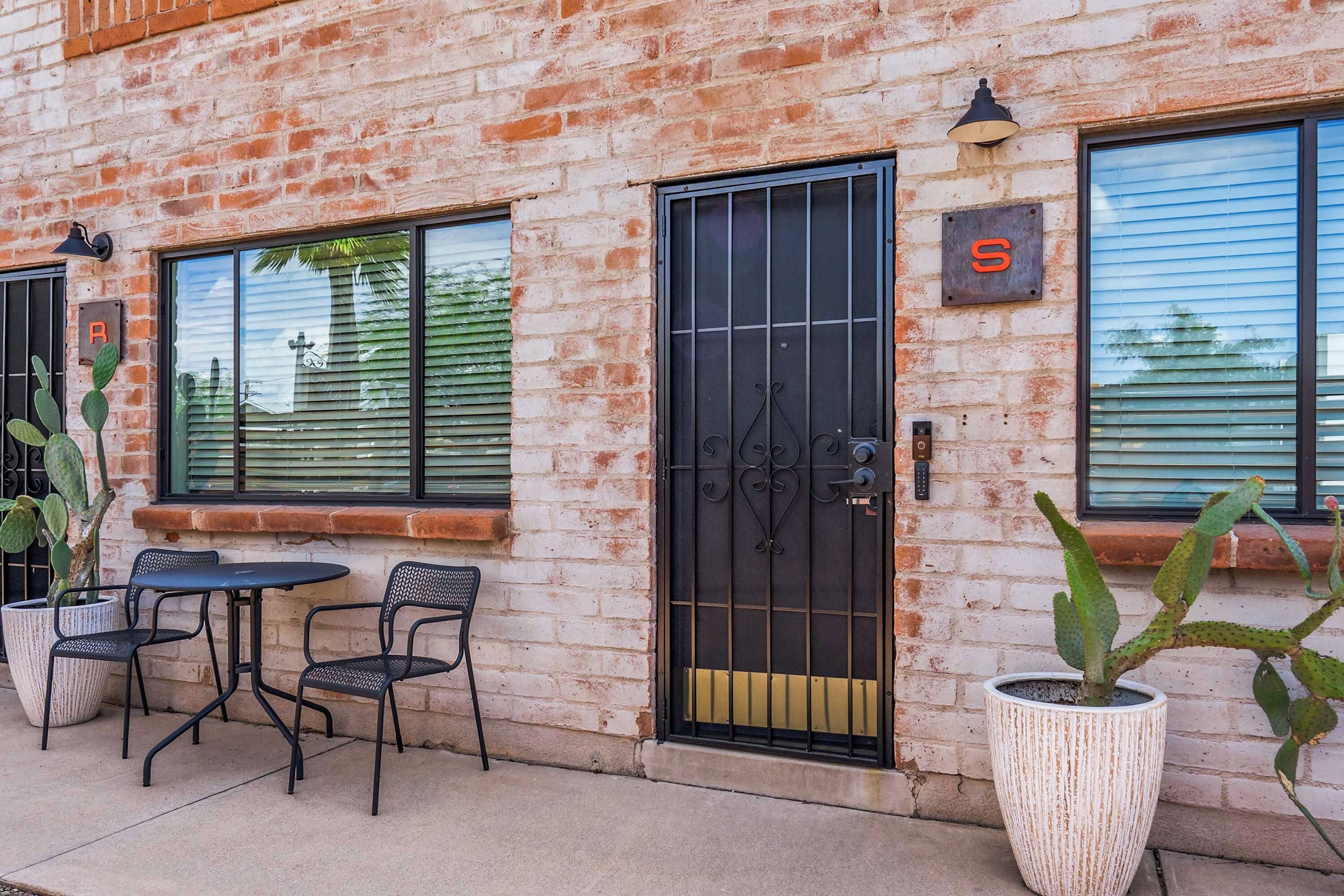 A brick building with a black metal gate, a small patio with two chairs, and a cactus plant.