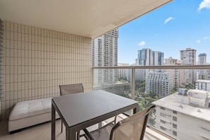 A balcony with a table and chairs, a cushioned bench, and a city view. - Cozy 1-bedroom condo with AC in Waikiki (Honolulu)