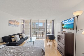 A modern living room with a sofa, a coffee table, a TV, and a balcony with a city view. - Cozy 1-bedroom condo with AC in Waikiki (Honolulu)
