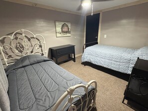 Quarto com estrutura de cama de metal, mesa de cabeceira, quadro na parede e piso coberto por carpete.