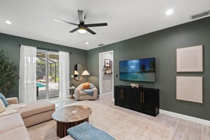 A modern living room with a flat-screen TV, a ceiling fan, and a sliding glass door leading to a pool area. - Heated Pool Getaway In Tampa Bay, Sleeps, Close to everything in Tampa Bay! (Tampa)