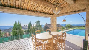 Terraço com mesa e cadeiras, vista para uma piscina e paisagem deslumbrante.