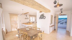 Uma sala de jantar com mesa e cadeiras de madeira, cozinha ao fundo e vista para o mar por uma porta aberta.