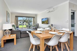 A modern dining area with a wooden table set for four, white chairs, and a television mounted on the wall. - Duck Pond Villa at Hunter Valley Golf Resort Views (Lovedale)