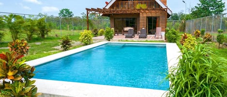 A swimming pool with a wooden deck and lounge chairs, surrounded by lush greenery.