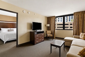 A hotel room with a bed, a TV on a dresser, a chair, a sofa, and a window with curtains. - Experience Vegas in Comfort • 1BR Suite • No Resort Fees! (Las Vegas)