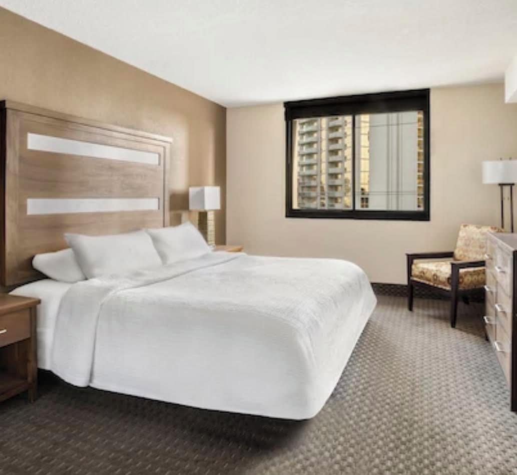 Ein Hotelzimmer mit einem großen Bett, einem Nachttisch, einem Sessel, einer Kommode und einem Fenster mit Blick auf Gebäude.