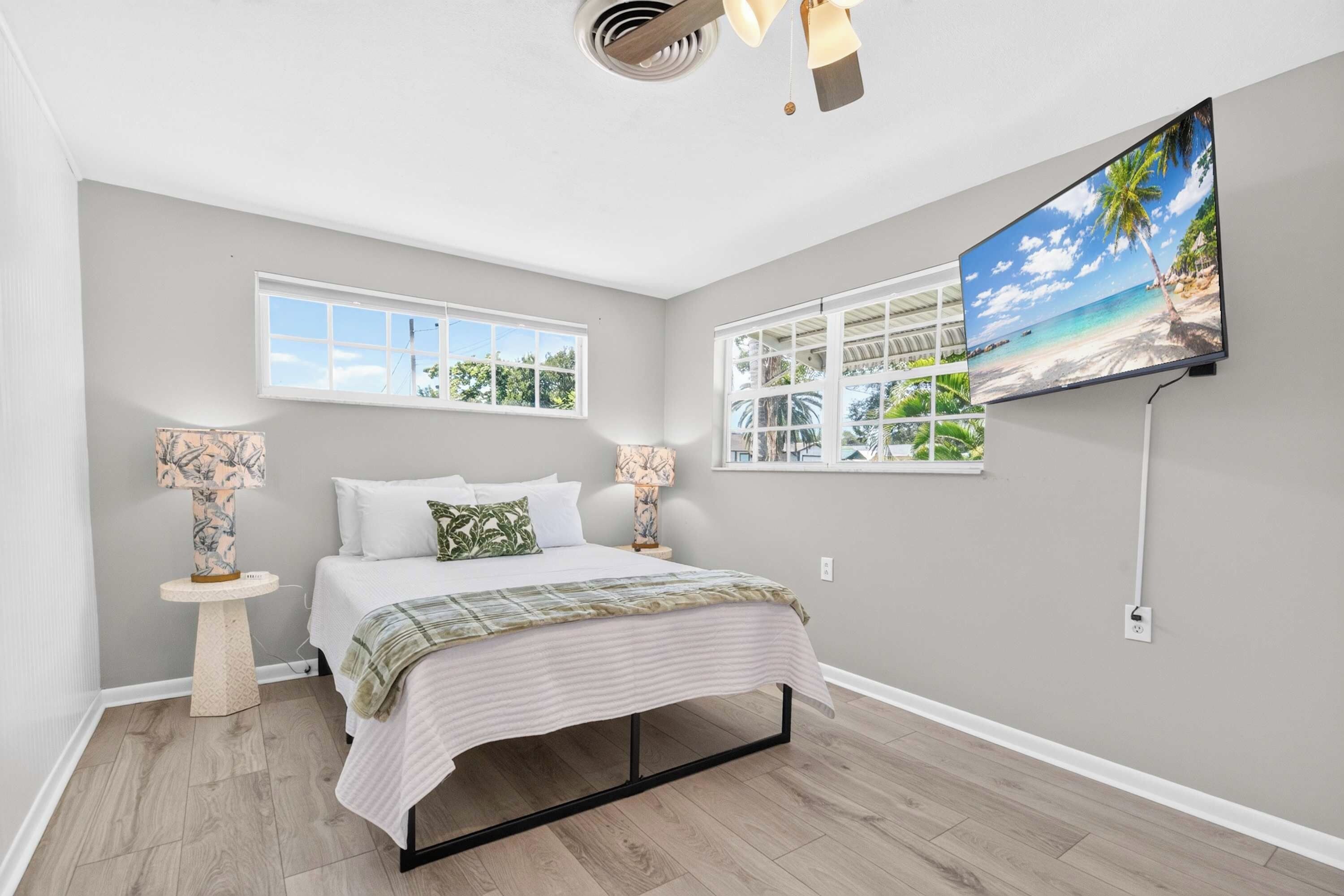 A bedroom with a bed, two bedside lamps, a ceiling fan, a TV on the wall, and two windows.