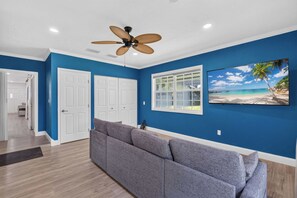 Ein Zimmer mit blauer Wand, Deckenventilator, Fenster mit Blick auf einen Strand und einem grauen Sofa.