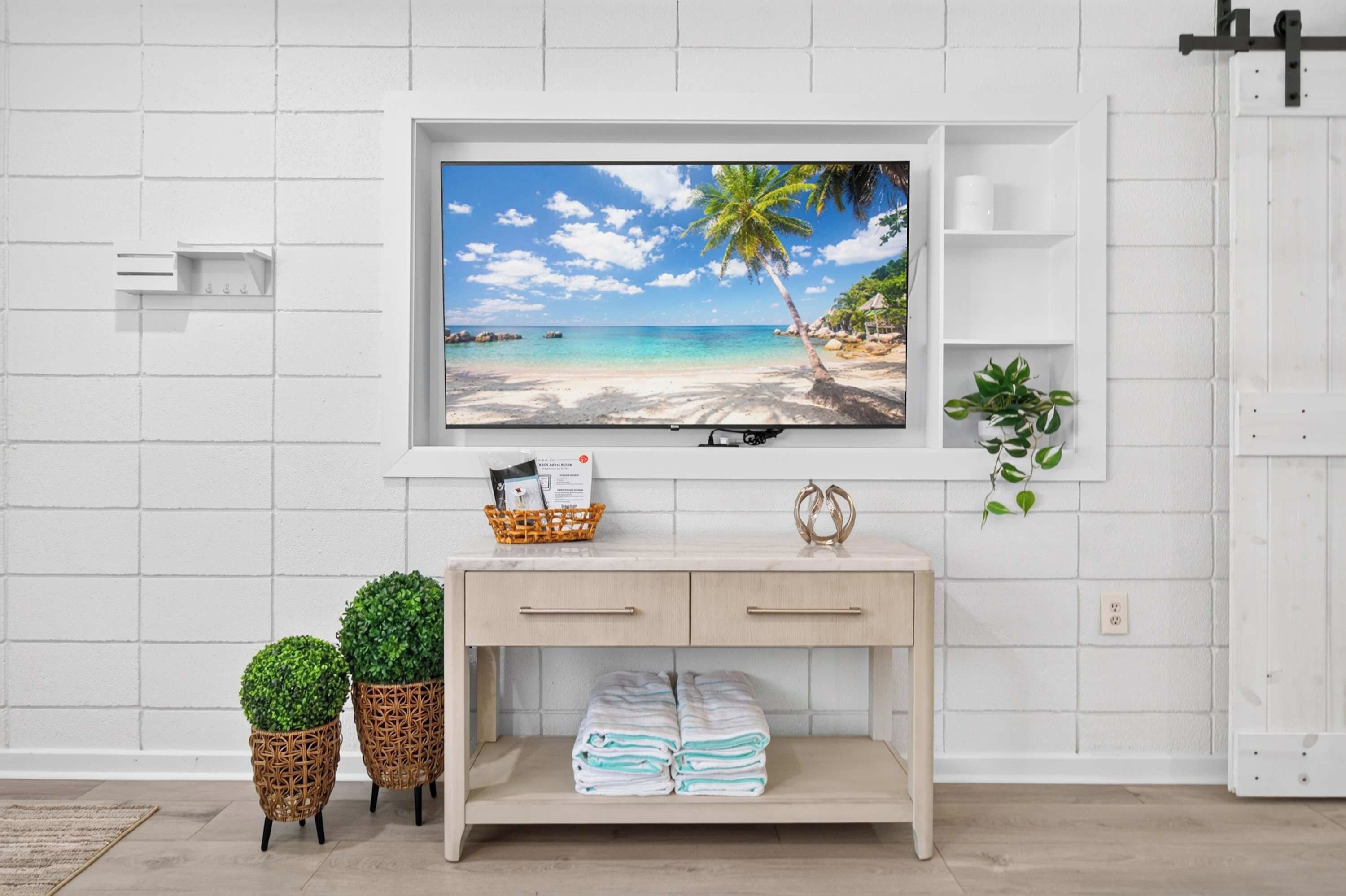 A modern living room with a TV mounted on a white brick wall, a wooden console table with a basket and folded towels, and two potted plants.