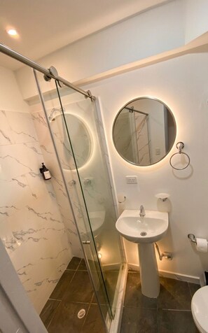 A bathroom with a glass shower enclosure, a pedestal sink, and a round mirror with built-in lighting. - Vrbo Property (Dosquebradas)