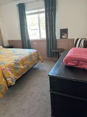 Ein Schlafzimmer mit einem Bett, einem kleinen Tisch, einem Stuhl, einem Fenster mit Vorhängen und einem gerahmten Bild an der Wand.