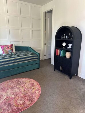 Ein Zimmer mit einem türkisfarbenen Sofa, einem runden Teppich, einem dunklen, gewölbten Bücherregal und einer weißen Tür.