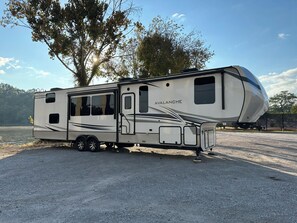 A white recreational vehicle with a curved design, labeled 'Avalanche,' parked on a gravel surface with trees in the background. - The Avalanche at River Rocks Landing (Gadsden)