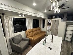 A compact living space with a sofa, a small armchair, a kitchen area with a sink, and a window with curtains. - The Avalanche at River Rocks Landing (Gadsden)