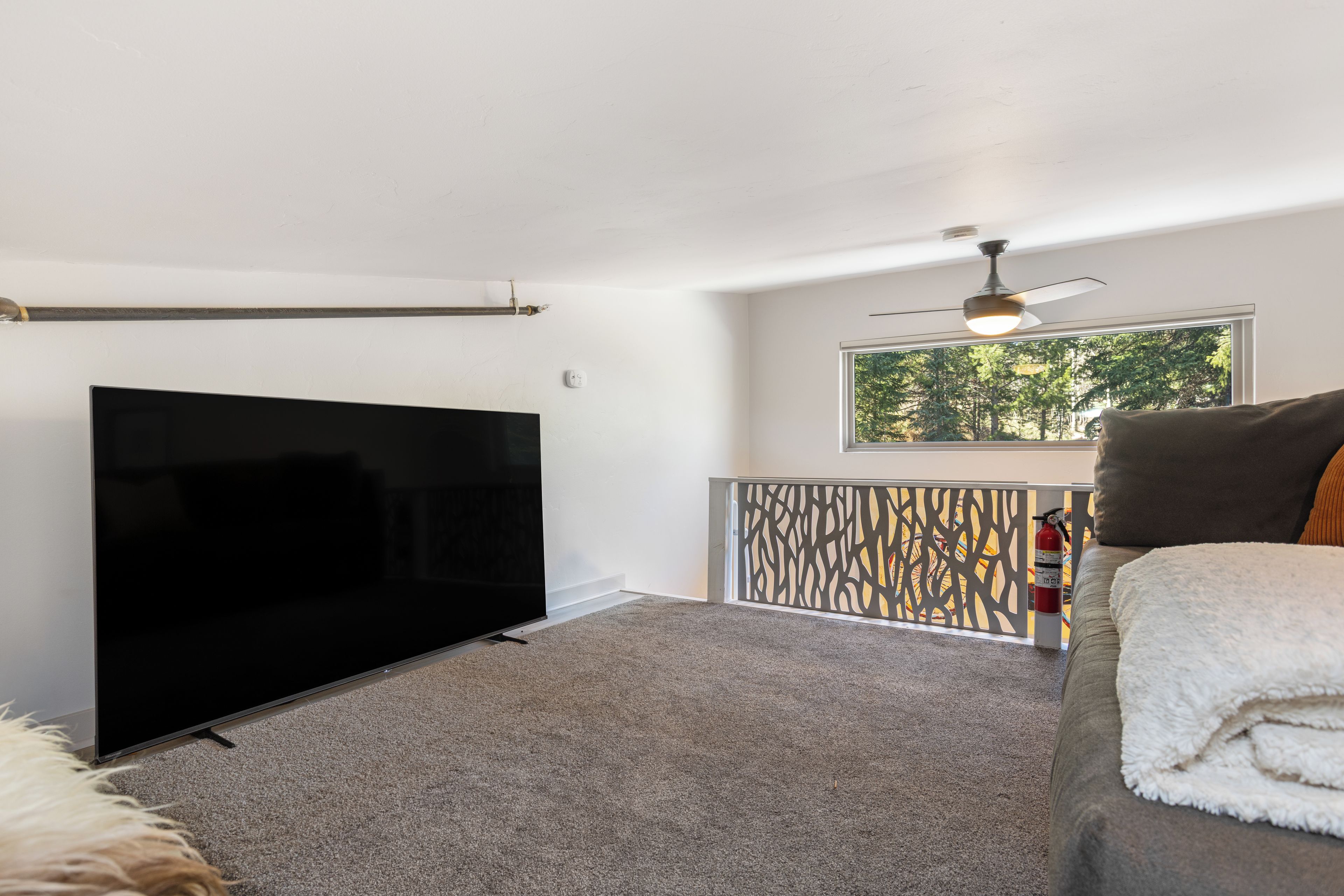 A living room with a flat-screen TV, a ceiling fan, a window with a view of trees, and a fire extinguisher.