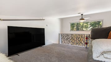 A living room with a flat-screen TV, a ceiling fan, a window with a view of trees, and a fire extinguisher.