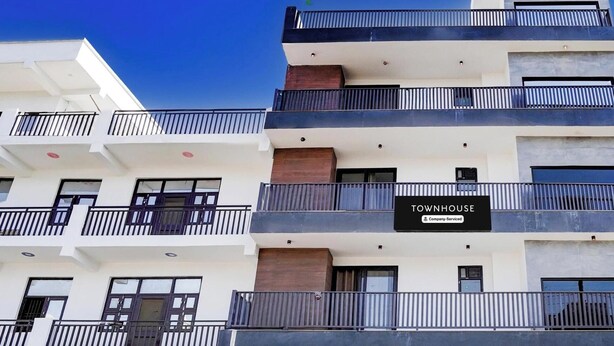 A multi-story residential building with balconies, a gated entrance, and a sign indicating it is a company-serviced townhouse. - Deluxe Townhouse Sector 143b (Noida)