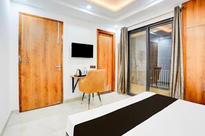 A hotel room with a bed, a desk with a chair, a television, and a sliding glass door leading to a balcony. - Deluxe Townhouse Sector 143b (Noida)