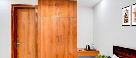 A bedroom with a wooden wardrobe, a bed, a bedside table with a kettle, and a framed picture on the wall.