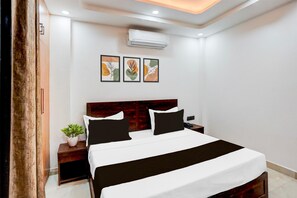 A hotel room with a bed, bedside tables, a nightstand, an air conditioning unit, and wall art. - Deluxe Townhouse Sector 143b (Noida)