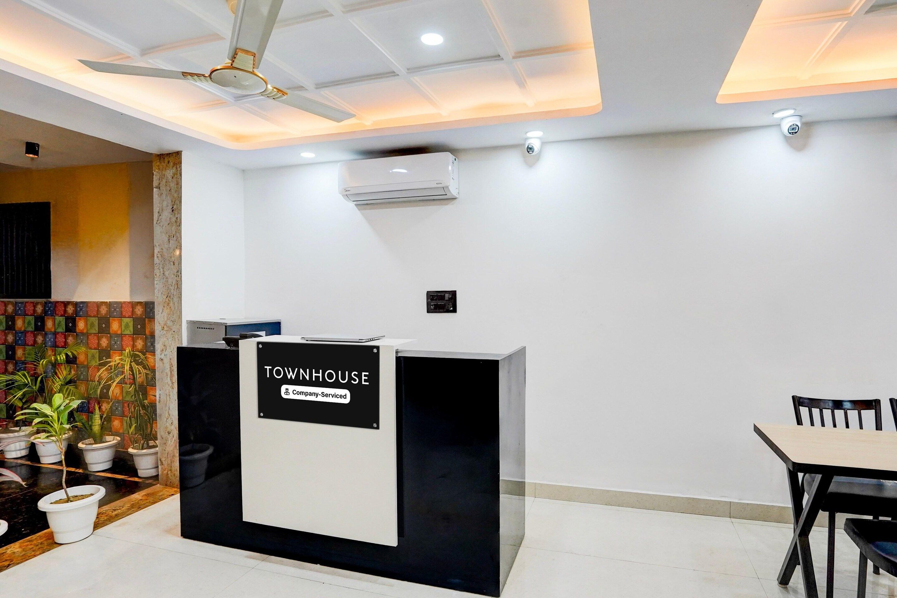 Un mostrador de recepción con la etiqueta 'TOWNHOUSE' y un cartel que indica 'Empresa de Servicios'. Hay un ventilador de techo, un equipo de aire acondicionado y una pared de azulejos con un diseño colorido.