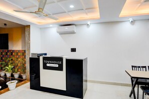 A reception desk labeled 'TOWNHOUSE' with a sign indicating 'Company Serviced.' There is a ceiling fan, an air conditioning unit, and a tiled wall with a colorful pattern. - Deluxe Townhouse Sector 143b (Noida)