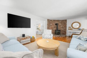 A living room with a fireplace, a round wooden coffee table, and a large flat-screen TV mounted on the wall. - Close to Mayflower Beach with deck, yard, and A/C. (Dennis)