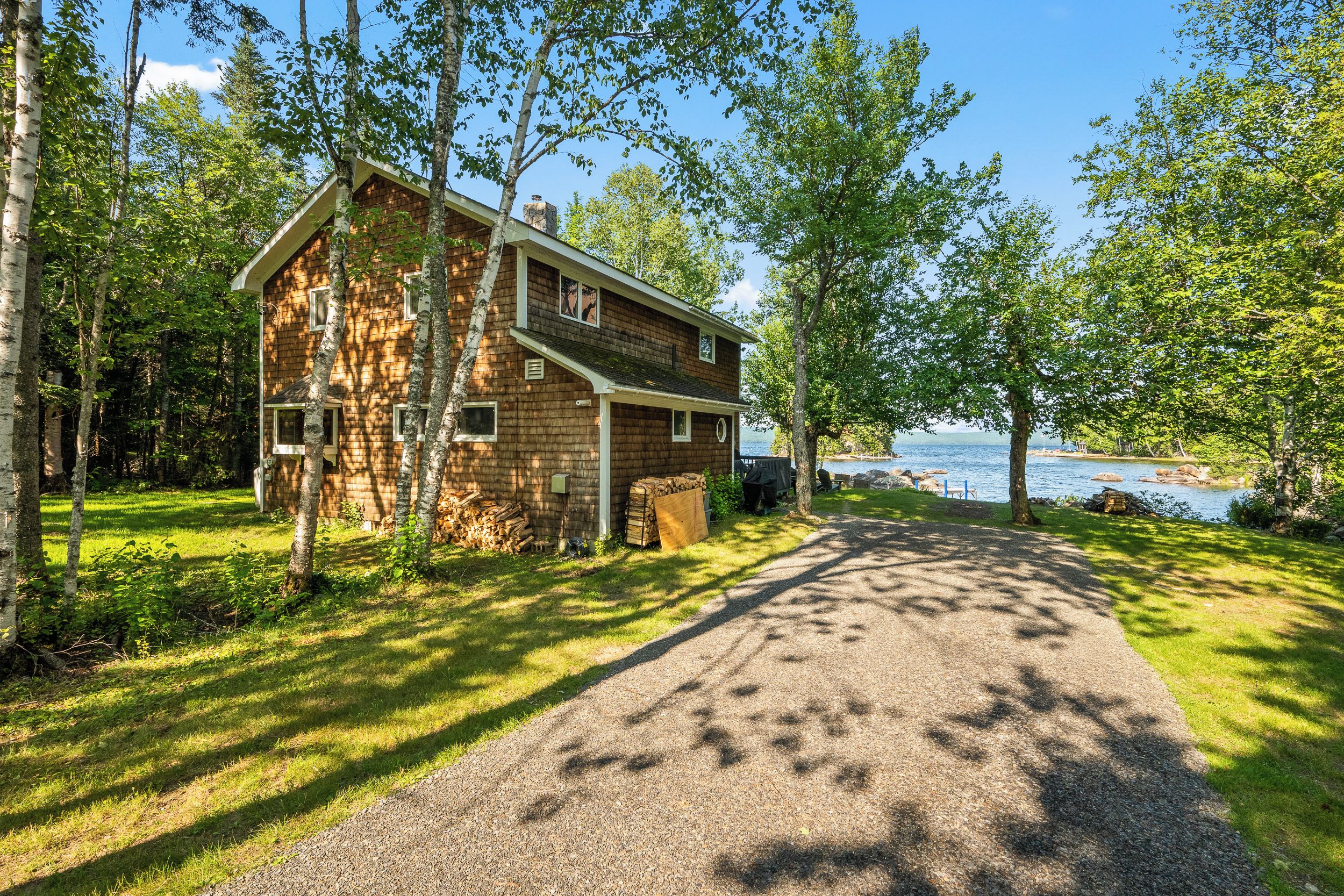 A two-story cabin with a gravel driveway leading to a dock over water.
