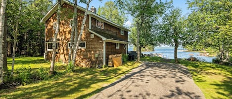 A two-story cabin with a gravel driveway leading to a dock over water.