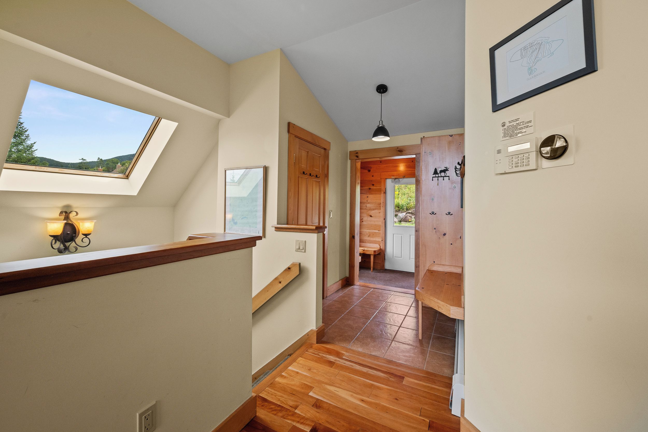A hallway with wooden flooring, a skylight, a framed picture, and a door leading to a room with a view of the outdoors.