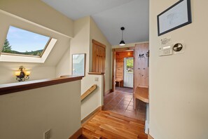 Un couloir doté d’un plancher en bois, d’une lucarne, d’un tableau encadré et d’une porte donnant sur une pièce avec vue sur l’extérieur.